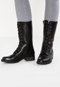 Botas de cuero negras hasta la rodilla con punta redonda, cordones en la parte frontal, cremallera lateral, acabado texturizado y tacón bajo en bloque.