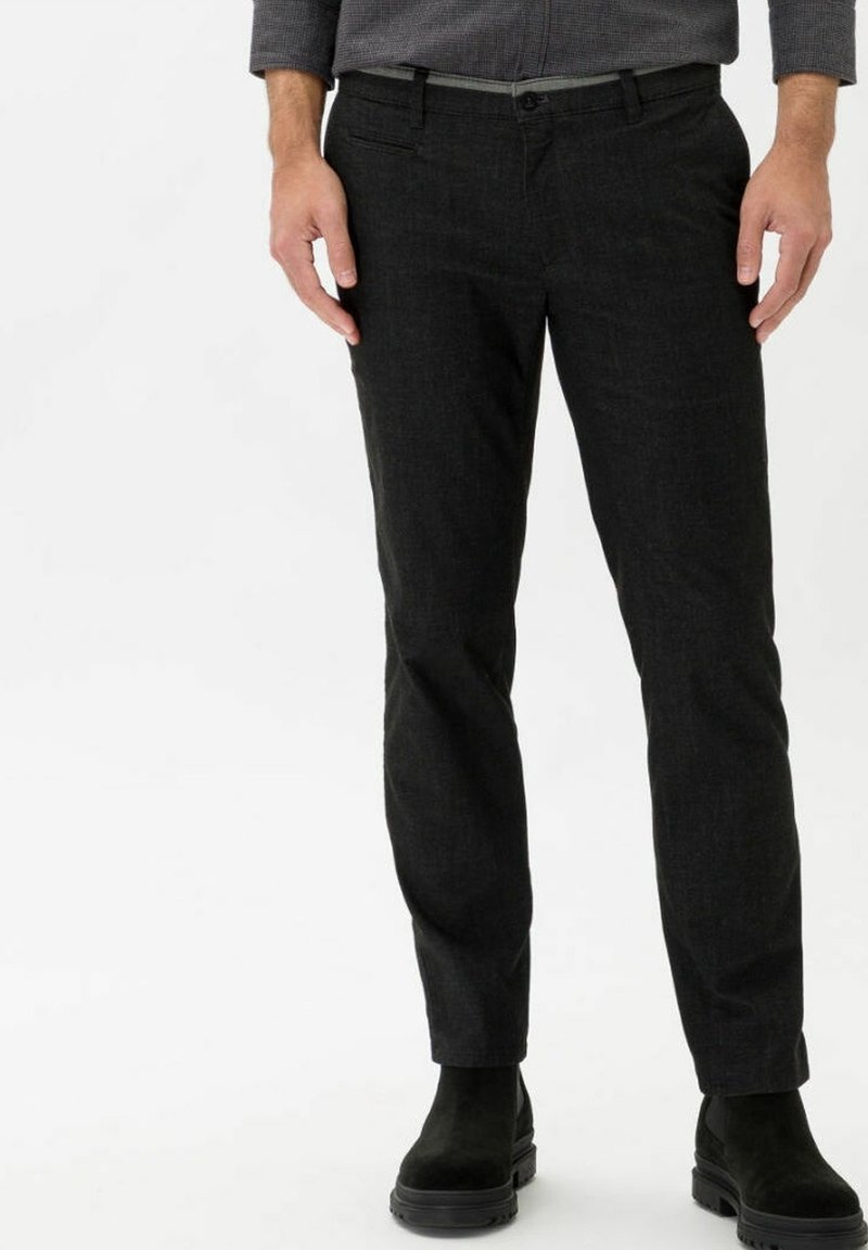 Homme portant un pantalon slim foncé et des bottines en daim noir, debout devant un fond blanc uni.
