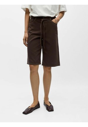 Personne portant un short marron foncé jusqu'aux genoux avec une ceinture nouée, une chemise blanche à manches courtes et des ballerines marron foncé à bout carré.