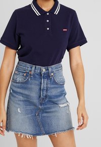 Femme portant un polo Levi's bleu marine rentré dans une jupe en denim bleu clair délavé avec ourlet effiloché et taille haute.