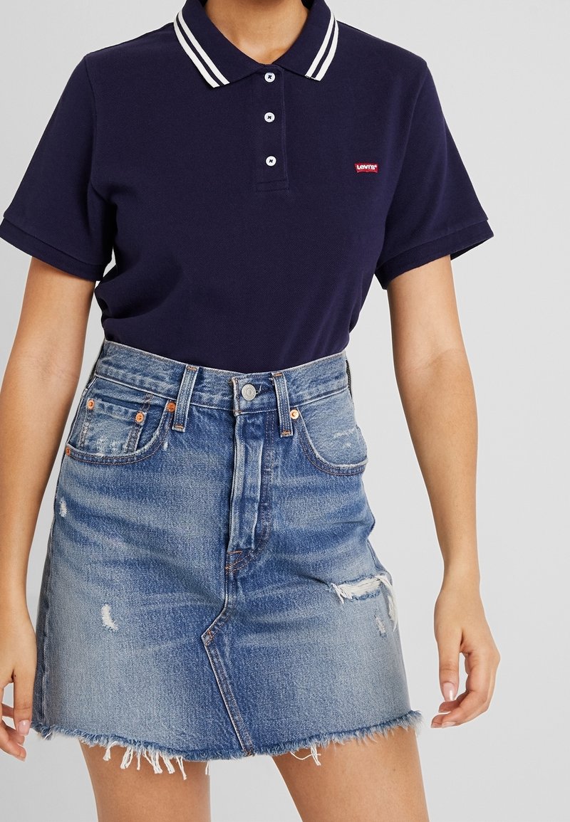 Femme portant un polo Levi's bleu marine rentré dans une jupe en denim bleu clair délavé avec ourlet effiloché et taille haute.