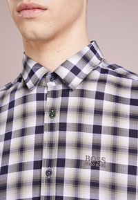 Zbliżenie na osobę w czarno-białą szaro-czarną w kratę koszulę Hugo Bossa, z widocznymi kołnierzykiem i guzikami.