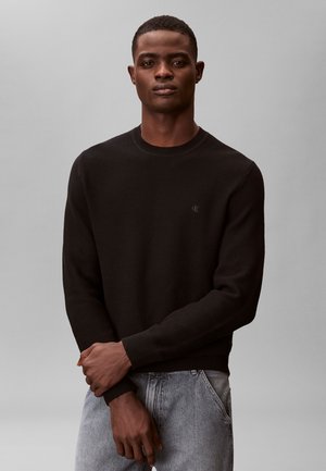 Bărbat purtând un pulover negru cu mâneci lungi și textură, și blugi gri deschis, stând cu mâinile încrucișate în față, pe un fundal simplu gri.