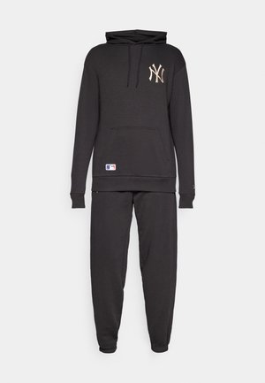Juodas medvilnės džemperis su priekine kišene ir reguliuojama kapišonu, turintis auksinį „Yankees“ logotipą. Derinamas su atitinkamomis siaurėjančiomis sportinėmis kelnėmis.