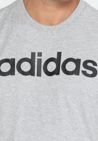 Šedé bavlněné tričko s velkým černým logem "adidas" na hrudi. Materiál vypadá hladce a má kulatý výstřih.
