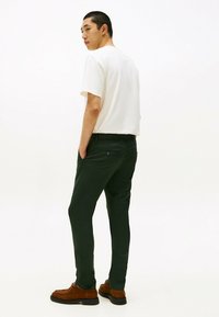 Pantaloni verdi realizzati in tessuto morbido con una vestibilità slim, dotati di tasche laterali e una tasca sul retro con bottone. Indossati con una maglietta bianca e scarpe marroni.