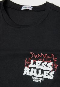 Czarny bawełniany t-shirt z okrągłym dekoltem, ozdobiony białą i czerwoną grafiką płomieni, na której widnieje napis "MNIEJ ZASAD" oraz "PRZYGODA CZKA".