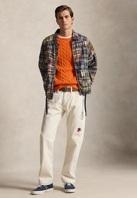 Mann iført en flerfarget lapperjakke, lys oransje kabelstrikket genser, hvite bukser med grafikk, brunt belte og blå joggesko som står med hendene i lommene.