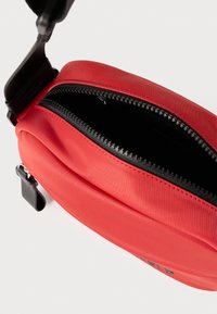Czerwona saszetka na zamek błyskawiczny o gładkiej teksturze; czarny zamek i uchwyt; kompaktowy kształt; logo z boku; częściowo otwarta, ukazująca wnętrze.