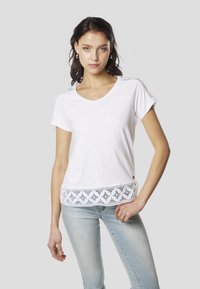 Biele bavlnené tričko s krátkymi rukávmi, výstrihom do V a prepracovanou čipkovanou lemovkou na spodnom okraji. Pod tričkom sú viditeľné svetlomodré rifle.