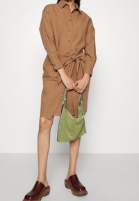 Abito marrone con bottoni e vita legata, abbinato a una borsa in suede verde e zoccoli in legno marrone. Texture liscia, design informale.