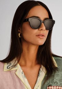 Γυναίκα με μακριά σκούρα μαλλιά που φοράει μαύρα υπερμεγέθη γυαλιά ηλίου, χρυσούς κρίκους σκουλαρίκια και πολύχρωμο πουκάμισο με παστέλ αποχρώσεις.