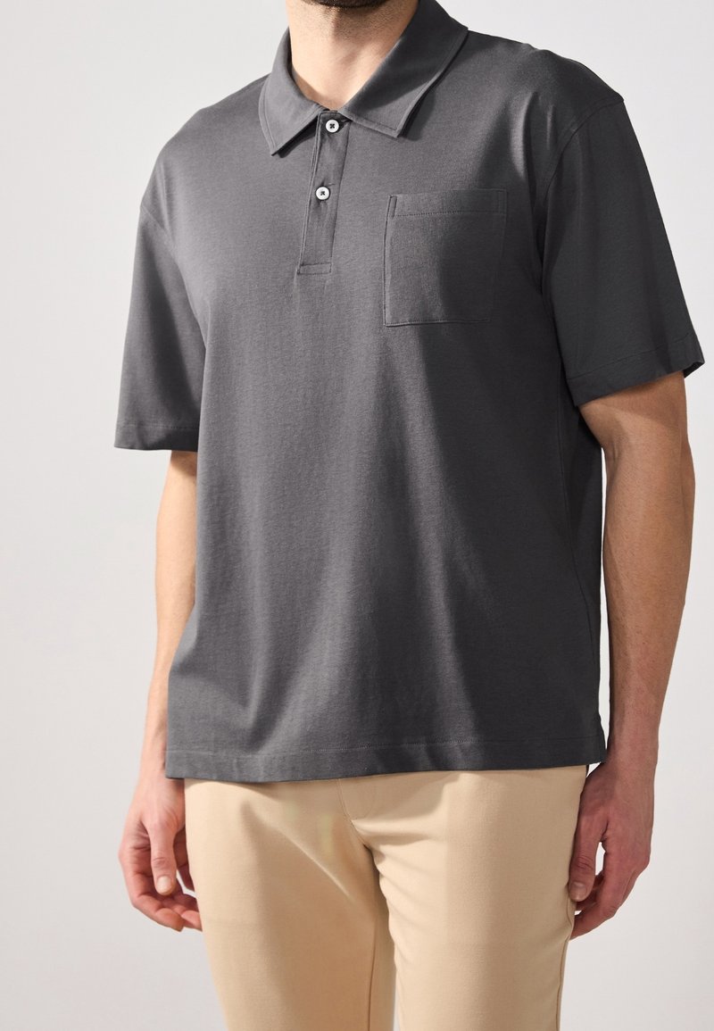 Homme portant un polo gris foncé à manches courtes avec boutons et poche poitrine, associé à un pantalon beige clair.