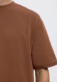 Nahaufnahme einer Person, die ein einfarbiges braunes Kurzarm-T-Shirt mit rundem Ausschnitt vor einem schlichten hellen Hintergrund trägt.