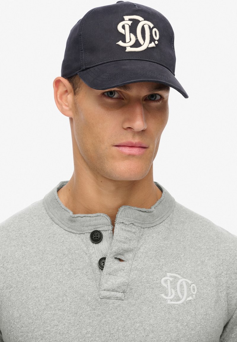 Lys grå henley skjorte med rund halsudskæring og to sorte knapper, prydet med et hvidt logo. Marineblå kasket med indbroderet logo foran.
