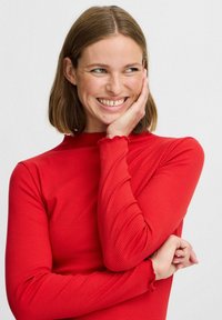 Rotes, geripptes Langarmoberteil mit hohem Halsausschnitt und gewellten Bündchen, aus einem dehnbaren Material gefertigt. Verfügt über eine taillierte Silhouette.