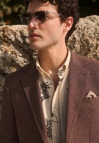 Blazer marron texturé sur une chemise crème avec des motifs de fougères noires. Des lunettes de soleil claires, un collier court et un pochette complètent le look.