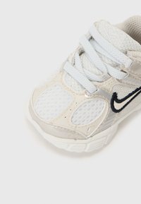 Chaussure de sport pour jeunes en blanc et gris clair, avec des panneaux en maille, un laçage et un logo swoosh bleu marine sur le côté. Semelle en caoutchouc.
