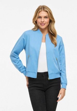 Femme portant une veste zippée bleu clair sur un haut blanc et un jean noir, debout avec une main sur la hanche, souriant légèrement.