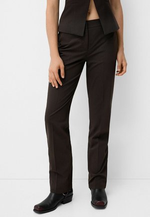 Frau trägt dunkel gestreifte gerade geschnittene Hosen, passende ärmellose Weste und schwarze Stiefeletten mit Absatz vor weißem Hintergrund.