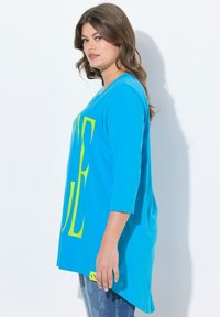 Blaues T-Shirt mit übergroßer neon-gelber Schrift, 3/4-Ärmeln und einem leicht verlängerten hinteren Saum, aus weichem Stoff.