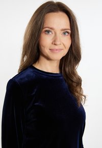 Top in velluto blu scuro con maniche lunghe e scollatura arrotondata, caratterizzato da una texture liscia e morbida. Capelli acconciati in onde morbide.