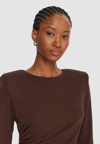 Top marrone a maniche lunghe con scollo arrotondato, caratterizzato da un tessuto morbido e lavorato e da un sottile drappeggio in vita. Dettaglio di orecchino a cerchio dorato.