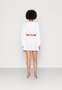 Conjunto em duas peças branco de eyelet, apresentando um top cropped com mangas longas e uma saia. O tecido é leve e possui um padrão texturizado.