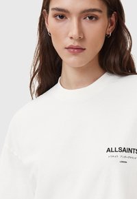 Biała bawełniana koszulka z okrągłym dekoltem, z czarnym napisem "ALLSAINTS" oraz akcentami w kilku językach na przodzie.