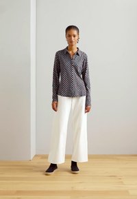 Schwarz-weiß gemusterte Hemdbluse kombiniert mit weißen weit geschnittenen Hosen und schwarzen Knöchelstiefeln an einem Modell mit geflochtenem Haar.