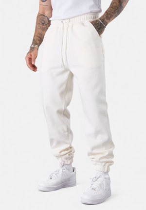 Pantalon de survêtement blanc avec taille élastique, poches latérales et poignets. Le tissu a une texture douce et lisse. Associé à des baskets blanches.