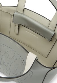 Sac fourre-tout en cuir gris avec des détails perforés, des poignées en gris clair contrastant et une poche intérieure. Texture lisse avec des bords noirs.