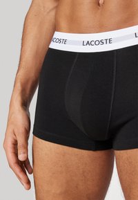 Schwarze Baumwoll-Boxershorts mit einem weißen Bund, auf dem "LACOSTE" in Schwarz gedruckt ist. Eine Nahaufnahme zeigt die Nähdetails und die Stoffstruktur.