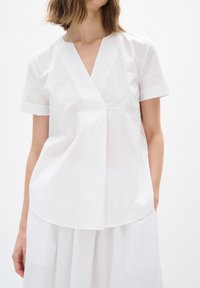 Top bianco a maniche corte con scollo a V e orlo ripiegato. Realizzato in un tessuto testurizzato, presenta un dettaglio a pieghe frontali e una vestibilità morbida.