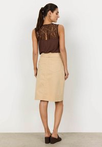 Haut sans manches marron avec un dos en dentelle, associé à une jupe beige de longueur genou dotée de poches frontales et d'une taille ceinturée. Chaussures compensées marron.