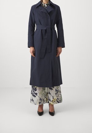 Mujer con un abrigo largo azul marino con cinturón, pantalones anchos con estampado floral y tacones negros de punta, posando frente a un fondo liso.