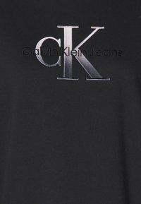 Czarna bawełniana koszulka z wyraźnym, srebrnym haftowanym logo "CK" oraz napisem "Calvin Klein Jeans" poniżej w eleganckiej czcionce.