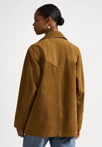 Sötét hajú, kontyba kötött hajú nő barna velúr dzsekit és kék farmert visel, hátulnézetből, egyszerű fehér háttér előtt.