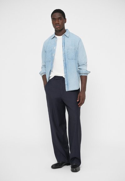 Hellblaues Jeanshemd über einem weißen T-Shirt, kombiniert mit dunkler Nadelstreifenhose. Schwarze Schuhe vervollständigen das Outfit. Einfaches Design und Farben.