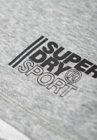 Textil gris con un logotipo negro prominente que dice "SUPERDRY SPORT." Incluye dos franjas negras paralelas encima del texto.