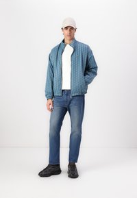 Blue patterned bomber jacket with a zip, worn over a white shirt, paired with blue jeans and black athletic shoes. Gray cap on the head.