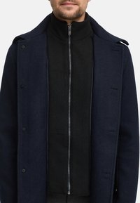 Marineblauer Wollmantel mit strukturiertem Kragen und Knöpfen, über einem schwarzen gerippten Reißverschluss-Pullover getragen, der ein modernes, maßgeschneidertes Design präsentiert.