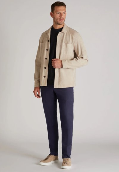 Veste beige clair avec des boutons marron, deux poches avant, associée à une chemise noire et un pantalon bleu marine. Présente des baskets beiges avec des semelles blanches.