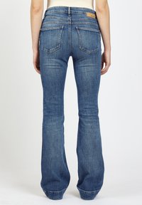 Frau in blauen Schlagjeans mit zwei Gesäßtaschen, die von hinten gegen einen weißen Hintergrund steht.