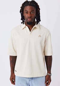 Polo shirt color crema a maniche corte con un motivo testurizzato, dotato di colletto e di un piccolo logo sul lato sinistro.