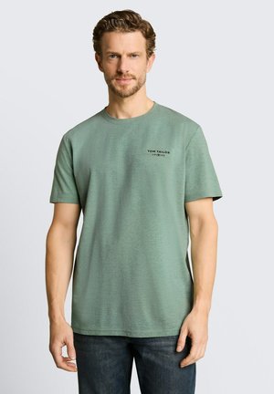 Mężczyzna z krótkimi kręconymi brązowymi włosami, ubrany w jasnozielony t-shirt Tom Tailor i ciemne dżinsy, stojący na jednolitym tle.