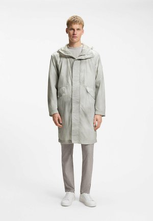 Joven con un impermeable largo gris claro con capucha, pantalones grises y zapatillas blancas, de pie sobre un fondo blanco y sencillo.