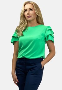 Zielona satynowa bluzka z falbaniastymi rękawami, okrągłym dekoltem i gładką teksturą, zestawiona z ciemnoniebieskimi, dopasowanymi spodniami z małym akcentem.