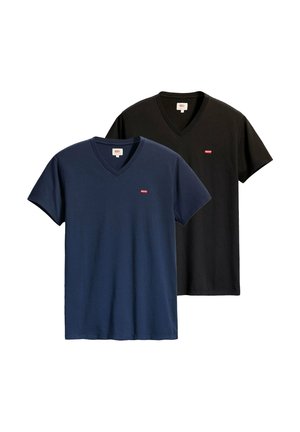 Junge Person mit welligem braunem Haar trägt ein schwarzes Levi’s T-Shirt mit V-Ausschnitt und hellblauen Jeans, steht vor weißem Hintergrund.