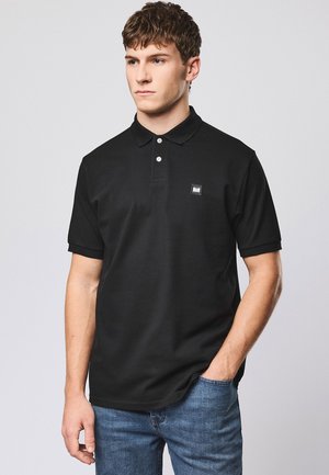 Joven con una camisa polo de manga corta negra con parche de logo, jeans azules, de pie con una mano en el bolsillo, mirando hacia la izquierda sobre un fondo gris claro.